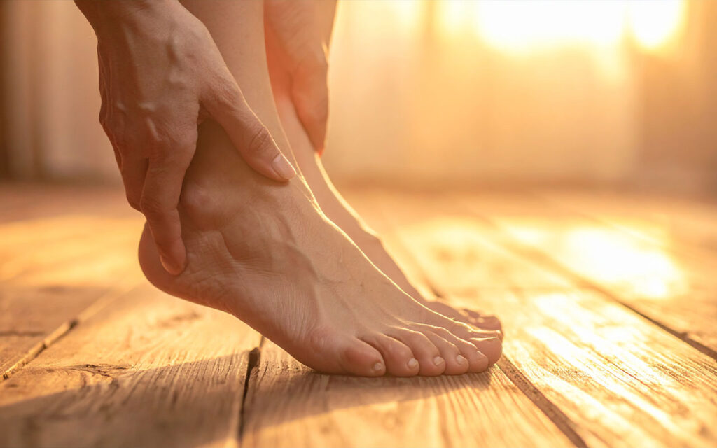 A pair of bare feet resting on a warm wooden surface, seen from above. One thumb gently presses the sole of the foot at the arch area. Warm, golden ambient light. Clean and calm aesthetic, soft focus background. Skin tones natural and healthy. No clinical setting — feels like a home wellness practice.