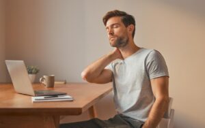 A man sitting at a desk with a laptop, eyes closed, hand pressing the back of his neck — illustrating the neck and shoulder tension that is a classic sign of Small Intestine Meridian chi stagnation in TCM